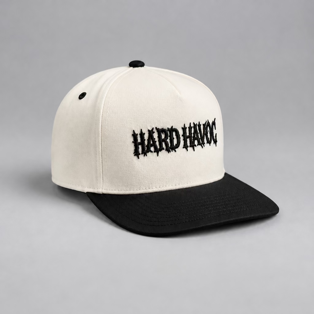 A off white and black baseball cap with 'HARDHAVOC' embroidered in black gothic-style lettering on the front, resting on a light wood surface against a plain light gray wall.