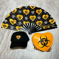 Black cap, black folding fan with biohazard symbols, and orange heart-shaped object with biohazard symbol on a gray background.