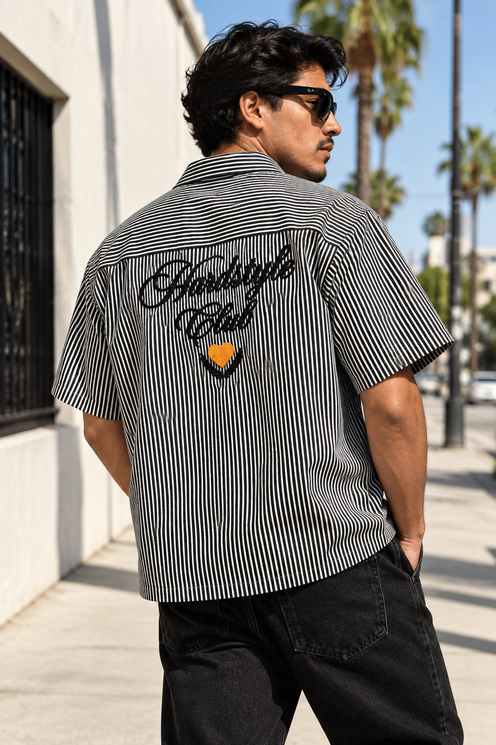 A short-sleeve button-up shirt with thin black and white vertical stripes, featuring 'Hardstyle Club' in black script and a small orange heart graphic on the back, laid flat on a gray fabric background.